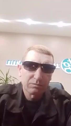 A man wearing sunglasses and a black shirt sits in front of a wall with a logo. He is looking directly at the camera. There is a plant on the left side of the image. The lighting suggests an indoor setting, possibly an office.