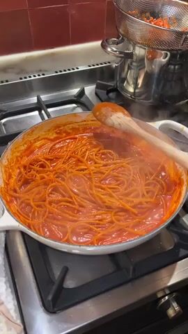 The video shows spaghetti with tomato sauce in a wide, white pan on a stainless steel stovetop. Steam rises from the pasta and sauce, indicating it's hot. A wooden spoon rests partially in the pan. In the background, a stainless steel pot with a strainer sits on the stovetop as well. The kitchen features a tiled backsplash.