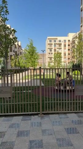A brief, static shot from behind a metal fence shows a playground area with three children on a merry-go-round. The children are partially obscured by the fence and the merry-go-round. The playground is in a courtyard surrounded by modern apartment buildings. The sky is clear and blue. There is a sign on the fence.