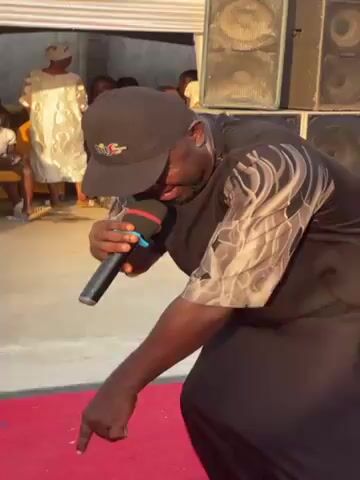 The video shows a person with dark skin singing into a microphone while pointing down. The person is wearing a black cap, a black shirt with a light gray pattern on the sleeves, and black pants. The person is standing on a red carpet in what appears to be an outdoor event setting. Several large speakers are stacked to the right of the person, and there are other people visible in the background. Decorations including white and yellow fabric are present. The person seems to be enjoying themself, smiling while singing.
