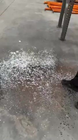 The video shows a warehouse setting with metal shelving, a man, and a pile of shiny debris on the floor. The man, wearing a black hooded sweatshirt with the word "STAFF" on it, smiles and looks at the camera. The shiny debris is scattered across the concrete floor. A few orange metal beams are visible on the shelves and floor around the debris.