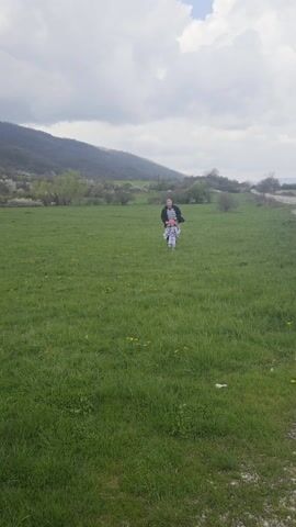 A man and a young girl are walking towards the camera in a grassy field. The man is wearing a striped shirt, dark pants, and a dark jacket. The girl is wearing a patterned outfit and a pink hat. The man briefly touches the girl's hat and then they both start running. The field is green with some yellow flowers, and there are trees and a mountain in the background. The sky is overcast.