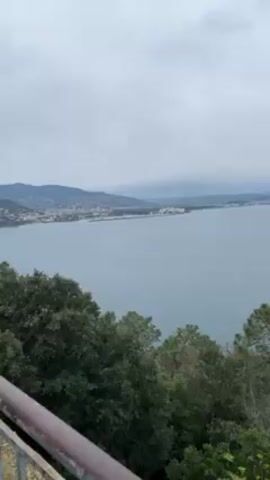 The video shows a view of a body of water with a coastline visible in the distance under an overcast sky. In the foreground, there are trees and a portion of a metal railing. The water is calm with a slight ripple on the surface. The distant coastline is lined with buildings, likely a city or town. The overall scene suggests a coastal viewpoint, possibly from a hill or elevated area.