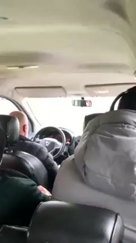 From the perspective of someone in the back seat of a car, we see the interior of the vehicle. A man is driving, and there are at least two other passengers. The windshield wipers are on, indicating rain. The car is driving on a road with green grass on either side.