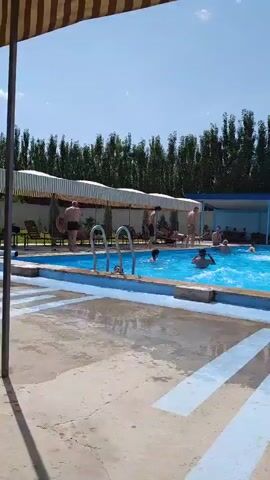 The video shows a group of people swimming and relaxing at an outdoor swimming pool on a sunny day. The pool is surrounded by a paved area with blue and white markings. There are lounge chairs and umbrellas around the pool. In the background, there are trees and buildings. Some people are swimming in the pool, while others are sitting by the side. The water in the pool is blue and clear. There are several people in the water, some of whom appear to be playing or splashing. The overall atmosphere seems to be one of leisure and recreation.