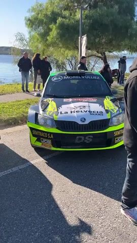 The video shows four race cars parked in a row on an asphalt road on a sunny day. In the background is a body of water with trees on the far side. At the edge of the water, several people are standing and watching the cars. The first car is black and yellow with the number 25 on the hood. The next car is white with the number 74 on the side. The third car is white. The fourth car is black with red accents and the number 25 on the side. The camera moves along the row of cars, showing them in detail.