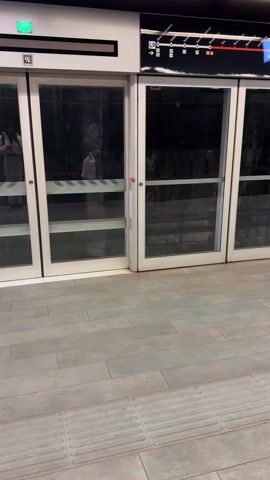 The camera pans across a modern subway platform with gray tile floors and a row of white sliding doors, typical of a platform screen door system. Behind the doors are visible the subway tracks. A digital display above the doors alternates between Spanish and English, announcing the next train’s destination as Aeroport T1. Passengers are visible on the platform, some standing near the doors and others sitting on benches. The ceiling is exposed, showing pipes and other infrastructure, with mounted screens displaying information.