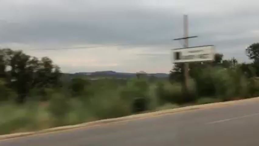 The video shows a view from a moving car. The road is on the right side of the frame, and beyond it, there's green vegetation with trees. In the distance, there's a range of hills under a cloudy sky. There are power lines visible in the landscape. The camera moves quickly, making the scenery appear blurry at times. Towards the end of the video, the edge of the car window frame is visible on the left side.