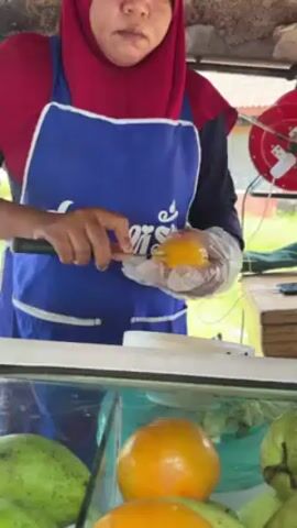 A woman wearing a red hijab and a blue apron with Thai text on it stands behind a white counter, preparing fruit. She has plastic gloves on her hands and uses a large knife to cut a yellow mango. She cuts off both ends and slices the sides into segments, placing them into a clear plastic bag. The counter is lined with a glass display case filled with green and orange fruits, likely mangoes and guavas. In the background, a red electrical cord reel is visible on the side of the cart. The environment appears to be outdoors, likely at a street vendor stall.