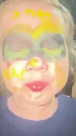 A young girl is captured in a close-up video. She is wearing dark navy blue clothing and has face paint in yellow, black, and green. The paint appears to be smeared in places. Above her eyes is a mask-like shape, with the word "пчела" written on her forehead, also in yellow paint. She has light skin, brown eyes, and visible teeth. The background suggests an indoor setting, possibly a hallway, with a door visible in the distance. At one point in the video, she extends her middle finger towards the camera. The girl seems to be speaking and animated.