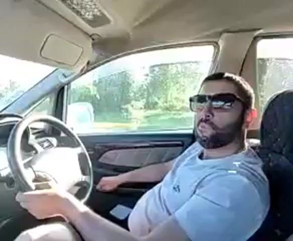 The video begins inside a car, showing the driver, a man with a beard and sunglasses, behind the steering wheel. The camera then shifts to the dashboard, looking forward. The car is on a highway, with another white car in front of it. Trees line the side of the road, and the sky is clear and blue. The video appears to be taken from a dashboard camera.