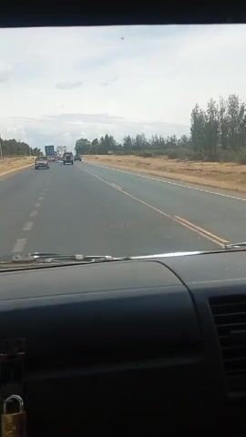 The video shows the view from the dashboard of a moving vehicle, likely a van or truck, driving on a highway. The dashboard is dark and textured, with air vents and a small padlock hanging from a bracket. Through the windshield, the road stretches ahead, divided by yellow lines. There are other vehicles on the road, including a car and a semi-truck in the distance. The sky is overcast, and the landscape on either side of the road consists of dry, grassy fields and scattered trees.