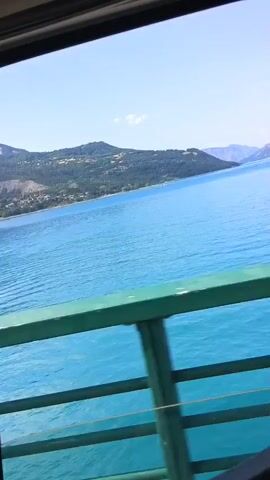 The video shows a view from a ferry looking out at a bright blue body of water. On the far side of the water, there is a coastline with green hills and some scattered buildings. The sky is clear and blue. A green railing is visible in the foreground, suggesting the camera is on a boat or ferry. The water appears calm with gentle ripples.
