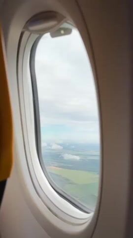 The video is a close-up, vertical shot through an airplane window. The window frame is white and oval-shaped, with a small silver latch visible at the top. The window's thick rubber seal is black and slightly curved inward. Outside the window, a vast landscape unfolds under a cloudy sky. The ground is a patchwork of green fields, some appearing cultivated with distinct patterns, interspersed with darker, possibly wooded areas. A few scattered white clouds drift through the blue sky, casting subtle shadows on the land below. The perspective suggests the airplane is at a moderate altitude, offering a wide, panoramic view of the terrain. The overall lighting is diffused, consistent with an overcast day.