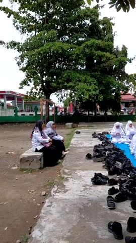 A group of individuals dressed in white robes and headscarves are seated on a blue mat in an outdoor setting. The individuals appear to be young, likely students, as they are seated attentively in rows, facing a direction out of frame. The setting is a courtyard or open space with buildings visible in the background. Several pairs of shoes and sandals are lined up along the side of the mat. Two women in the front left are sitting on a concrete block, observing the group. There is a large tree in the foreground. The weather appears to be overcast or cloudy.