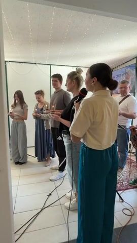 A group of seven people is gathered in a well-lit room with a festive atmosphere. Six individuals stand in a line, each holding a small plate with a lit candle. A woman in teal pants stands slightly forward, holding a microphone and appearing to speak or sing. Behind her and to the right, a man plays a bass guitar. The room has white tile flooring, and the walls are decorated with string lights. There's a screen in the background displaying text in a different language, along with a picture of snowy mountains. The people are dressed casually in clothing like jeans, t-shirts, dresses, and sneakers. The setting suggests a small gathering or event, possibly religious or celebratory in nature.