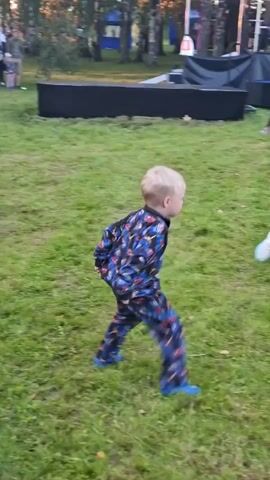 The video shows an outdoor event with several people of various ages gathered on a grassy area. In the foreground, a young boy with blond hair is wearing a blue pajama-like outfit with a colorful pattern and blue shoes. He is seen dancing, with his arms extended upwards and his legs apart, suggesting he is jumping or in mid-dance. Other people are visible in the background, some of whom also appear to be dancing or interacting. The environment suggests a casual gathering, possibly a festival or an outdoor party, with some structures or tents in the distance. The lighting indicates it is likely late afternoon or early evening.