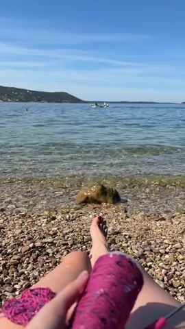 The video shows a person's legs and hands as they crochet by the sea. The person is sitting on a pebble beach with their legs extended toward the water. In their right hand, they hold a skein of pink yarn with sequins. Their left hand is engaged in crocheting a pink item with a crochet hook. The sea is clear and calm, with small waves lapping at the shore. In the background, there are several people in the water, some on paddleboards, and a hilly coastline. A pigeon walks on the pebbles in front of the person. The sky is blue with a few white clouds.
