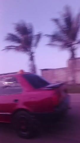 The video shows a view from a moving vehicle, likely a car. The camera is positioned looking out the side window. The scenery is coastal, with buildings, palm trees, and the ocean visible. The colors in the video have a purplish tint, possibly due to lighting or a filter. The car appears to be driving along a road that runs parallel to the coastline. The focus is on the passing landscape, which includes beachside structures and glimpses of the ocean waves. The speed of the vehicle causes motion blur in the video.