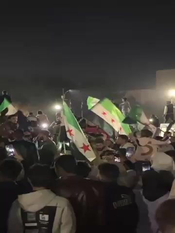 At night, a gathering of people are waving green and white flags with red stars, some are standing on an elevated platform, and some are recording the scene with their phones.