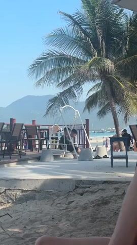 The video shows a sunny day at a beach resort. In the foreground, a person's legs are visible, partially obscuring the sandy beach. In the middle ground, a white swing set is occupied by two individuals. One person is sitting and appears to be talking or interacting with the other, who is also seated. Behind the swing set, a palm tree sways gently, with its fronds reaching towards the clear blue sky. In the background, there is a body of water, possibly the ocean, with a coastline and mountains visible in the distance. There are also tables and chairs set up on a wooden deck, suggesting an outdoor dining or seating area. Another person can be seen in the background, to the right of the swing set, near a blue chair. The overall atmosphere is relaxed and tropical.