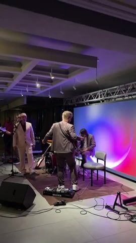 A band is performing on a stage with a large LED screen in the background displaying abstract, colorful visuals. The screen shows a pink and blue gradient with concentric circles radiating outwards. The stage lighting is purple and white.

On the stage, there are four musicians. In the foreground, to the left, a man in a light pink suit and matching headwear stands at a microphone, singing. He is wearing white sneakers. To his right, another man in a darker grey suit faces away from the camera, holding a guitar and looking at his phone. He is also wearing white sneakers and has a pedalboard in front of him. In the middle of the stage, behind the singers, is a red drum set. Another musician, wearing a dark suit and glasses, is seated at the drums, partially obscured. Further back and to the left, another person is visible playing an instrument, likely a guitar, but they are out of focus.

The setting appears to be an indoor venue, possibly a concert hall or event space, with a textured ceiling and modern light fixtures. There are speakers and cables visible on the floor, indicating a live performance. The overall mood seems energetic and focused on the music. The visible text on the pedalboard reads "MOOGER". The time of day is likely evening or night due to the stage lighting and indoor setting.