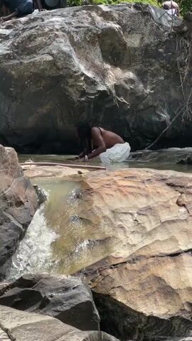 A person is crouched in shallow water near a rocky waterfall. The person is wearing a dark top and white shorts. They are reaching into the water with their hands. The water is clear and appears to be flowing over rocks. The surrounding area is rocky and covered with green vegetation. In the background, there are large boulders and trees. The sunlight suggests it is daytime. The overall scene appears to be a natural outdoor setting.