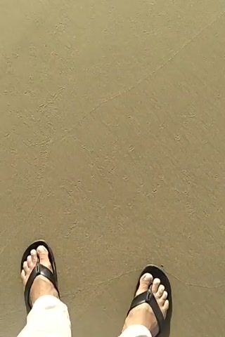 The video begins with a first-person perspective of someone standing on a sandy beach, wearing flip-flops. The camera pans up to reveal the ocean with gentle waves breaking on the shore. The sun is high in the sky, casting a bright reflection on the water and the wet sand. The overall scene is a peaceful view of a beach on a sunny day.