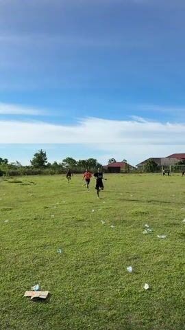 This media file captures an outdoor scene on a bright, sunny day with a clear blue sky dotted with wispy white clouds. The setting appears to be a grassy field, possibly a sports field, with some scattered debris including plastic bottles and a piece of cardboard. In the background, there are trees and a few buildings with red roofs, suggesting a rural or semi-rural location. There's also a goalpost visible on the right side of the frame.

The primary subjects are a group of young people, appearing to be males, engaged in running activities. Several individuals are in motion across the field, with one person in the foreground on the right side of the frame turning to face the camera. The general atmosphere seems casual and active, with the participants focused on their running. The bright sunlight and clear sky indicate it is daytime, likely late morning or early afternoon. The presence of the scattered debris might suggest a lack of consistent upkeep for the field. There is no visible text in the media file.
