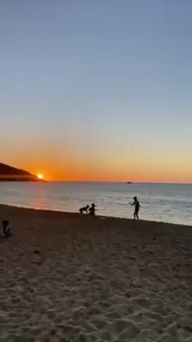 The video shows a beach at sunset. The sun is setting over the water, partially hidden behind a silhouetted hillside. There are several people on the beach, including a group of young adults or teenagers standing in a cluster, and several younger children playing in the sand and near the water's edge. The sky is clear and orange where the sun is setting, grading into a light blue further overhead. The sand is beige and appears slightly wet. Small waves are visible lapping at the shore.