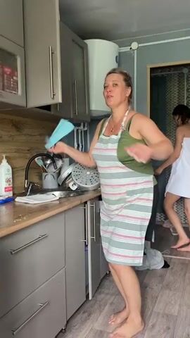 A woman in an apron stands in a kitchen, holding a blue sponge and singing. She is barefoot and appears to be dancing or performing. Behind her, another woman wrapped in a white towel walks away from the camera. The kitchen features gray cabinets, a countertop with dishes and cleaning supplies, a washing machine, and a water heater on the wall. The floor is wooden.