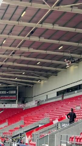 The video shows a person in military camouflage suspended from a zip line inside a stadium. They glide down the line toward the ground, where three or four other people in similar camouflage uniforms are positioned. The stadium has red seats and advertisements on the walls. The sky above is overcast.