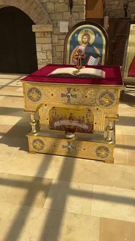The video opens with a close-up of a richly decorated golden altar with a red velvet cloth on top, upon which rests an open book and a cross. The altar is ornately designed with intricate patterns and features a depiction of the Last Supper on its front. In the background, a large icon of Jesus Christ is visible, set against a stone wall. As the camera pans to the right, more icons are revealed, including one of the Virgin Mary and another of a male saint. A young boy walks into the frame, briefly obscuring the icons before moving out of view. The camera then focuses on a series of large, framed religious icons, some of which are displayed on easels. The environment appears to be a church or a religious hall, with stone walls and a tiled floor. The lighting suggests it is daytime.