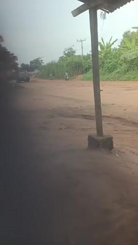 The video depicts a dirt road scene in what appears to be a rural area. The foreground is partially obscured by shadow, possibly from a structure or awning. In the middle ground, a dusty, reddish-brown dirt road is visible with vegetation lining the right side of the frame, including tall grass and banana trees. A wooden pole supports an overhanging structure. A blue van approaches, moving from the distance toward the camera on the dirt road. The sky is overcast, suggesting hazy or misty conditions.