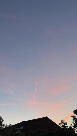 The video shows a time-lapse of a sky with pink contrails against a dusky purple backdrop. The contrails are from airplanes, leaving streaks across the sky. In the foreground, silhouetted against the sky, are the rooftops of houses and some trees. The scene is peaceful and serene, suggesting a sunset or sunrise. The camera is static, and the video is shot in portrait mode.