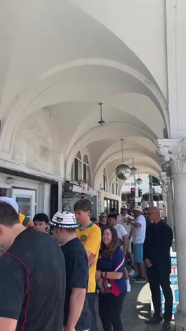 The video begins by showing a line of people under a white arched walkway. The camera pans to a storefront labeled "Venice Beach Mart" which sells groceries, beverages, snacks, tobacco, sandwiches, and deli items. The camera then shows a "No Stopping Anytime" sign with a number for impounded vehicles. Finally, the video shows a long line of people waiting outside a building with a blue and white facade. The building has large posters on its side with numbers and names. The line is cordoned off with stanchions.