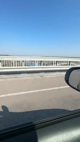 This video shows a view from inside a vehicle, looking out the passenger side window. The vehicle is traveling on a road, likely a bridge, given the railing and the expanse of water visible beyond it. The railing is white with metal bars and has lights or poles spaced along it. A tall, gray pole rises vertically on the left side of the frame. The road surface is asphalt with white lane markings. The sky is clear and blue, suggesting a sunny day. A shadow of the vehicle or its occupants is visible on the road in the foreground. The video is handheld and slightly shaky, indicating movement.