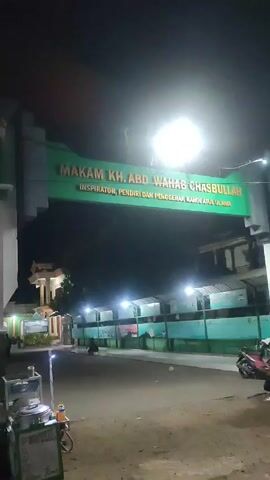 Here is a description of the scene in the video:

The video shows the entrance to the "Makam KH. ABD WAHAB CHASBULLAH" which is illuminated by several street lights at night. A food cart labeled "PENTOL" is visible on the left side. A few people can be seen in the background. A motorcycle is parked on the right. The scene is set outdoors, and the overall atmosphere is calm and quiet.