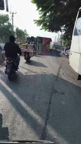 The video shows a traffic scene from the perspective of a motorcycle rider. In the foreground, the handlebars and instrument panel of a motorcycle are visible. The road ahead is paved with asphalt and appears to be somewhat rough.

Further down the road, several vehicles and people are present. A white scooter with a rider wearing a blue jacket and a white helmet is in the center of the frame, moving away from the camera. Behind it, a large green and red truck is overturned, its cargo spilling onto the road. The truck is partially covered by a red tarp. Several people, some wearing yellow vests and hard hats, appear to be involved in dealing with the situation. One person is standing near the overturned truck, seemingly directing traffic or assessing the scene.

On the right side of the road, another truck is visible, and more people in yellow vests are present. The environment appears to be an outdoor, possibly semi-urban or rural setting, with trees lining the right side of the road. The lighting suggests it is daytime, with sunlight casting shadows on the road. The overall impression is of a traffic incident or a road obstruction that is being managed.