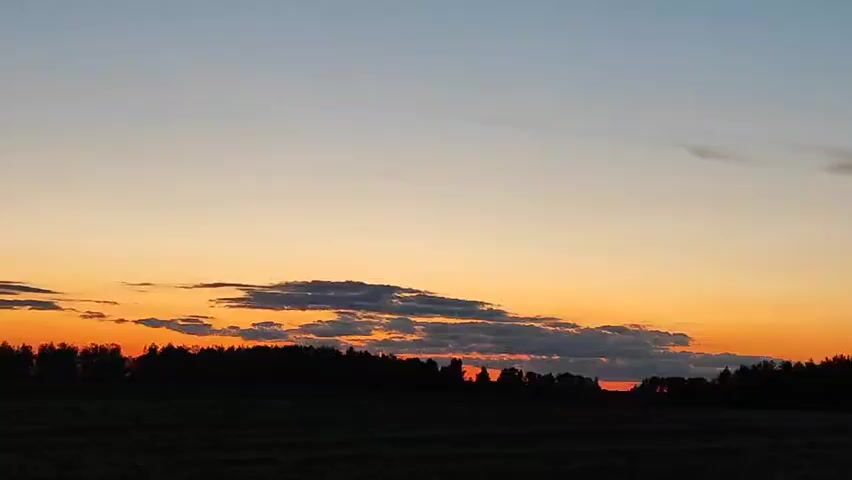 A wide shot captures a silhouetted treeline against a vibrant sunset sky. The sky transitions from a soft blue at the top to a warm orange and red near the horizon, with scattered clouds adding texture. The foreground is a dark, indistinct field, likely in silhouette due to the low light conditions. The overall mood is serene and peaceful.