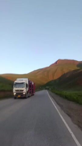 A highway runs through a mountainous landscape. The sun is low in the sky, casting long shadows across the rolling hills, which are covered in green grass and patches of reddish-brown earth. The road is paved and has a white line marking the lane. On the left side of the road, a car carrier truck is visible, its trailer loaded with vehicles. The sky is a clear, pale blue. The camera appears to be mounted on a vehicle moving along the highway.