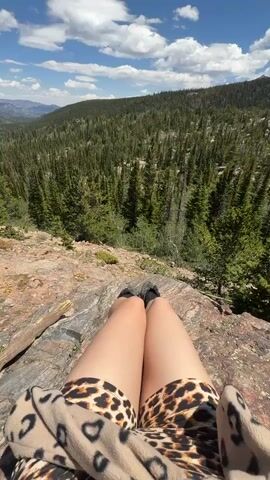 The video shows a first-person perspective of someone sitting on a rocky outcrop, looking out at a vast, forested mountain landscape under a bright blue sky with scattered white clouds. The person's legs are visible in the foreground, wearing leopard-print shorts and dark shoes. They are wearing a patterned fuzzy jacket or blanket over their legs. The ground beneath them is covered in large, reddish-brown rocks. The surrounding environment is dense with green pine trees covering the slopes of the mountains. In the distance, more mountains are visible. The camera pans slightly to the right, revealing more of the expansive valley and distant mountain range. The overall impression is one of peaceful solitude and appreciation of nature.