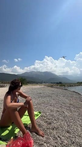 A woman in a bikini sits on a pebble beach, facing left, with her legs crossed. She wears sunglasses and has her arms resting on her knees. Behind her, mountains rise against a blue sky with scattered clouds. An airplane is visible in the sky. To the right, the sea meets the shore. In the foreground, a green towel and a red bag are on the ground.