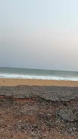 The video shows a beach with a man walking along the shore. The beach has a section of dark-colored, pebbly ground near the foreground. This transitions into a sandy area nearer the water. The sea is visible with small waves. A man in dark clothing is seen walking on the sand towards the right side of the frame. The sky is a pale blue color transitioning to a faint pink-orange hue near the horizon. The overall scene is peaceful and serene.