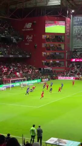 The video captures a soccer match from a spectator's perspective inside a stadium packed with fans, predominantly wearing red, reflecting the team colors. The stadium features a large, red structure with multiple viewing levels, adorned with logos from sponsors like "new balance" and "Red Cola." A large screen displays the ongoing game.

The field is green and well-lit, showing players in red and blue uniforms actively engaged in the game near one of the goals. The red team is on the attack. A player in blue makes a diving save near the goal. A couple of individuals wearing green vests stand near the camera's position, appearing to be field staff. The environment is vibrant with a palpable sense of excitement from the crowd.