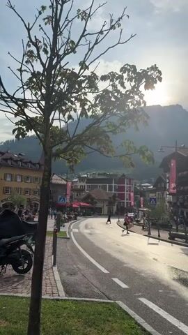 The video opens with a view of a tree with green leaves in the foreground. The sun is setting behind a mountain range, casting a warm glow over a town. The town features buildings with traditional architecture, including balconies adorned with flowers. A street runs through the town, wet from recent rain, reflecting the sunlight. Several people are walking along the street and sidewalks, some appearing to be tourists. There is a motorcycle parked on the left side of the street. The overall atmosphere is peaceful and idyllic.