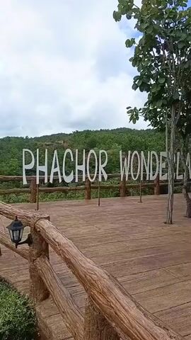 The video pans across a scenic overlook. In the foreground, a wooden railing with a rustic, log-like appearance surrounds a wooden deck. A black lantern is mounted on the railing. Large, white, block letters spelling out "PHACHOR WONDE" are positioned on stands behind the railing, also on the deck. Beyond the deck, the view opens up to a vast expanse of lush green forest and rolling hills under a cloudy sky. The camera moves from left to right, revealing more of the landscape and the scale of the forest.