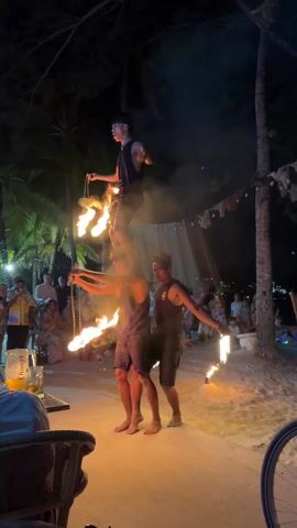 At night on a sandy beach, three men participate in a fire performance for an audience. The two men in the bottom layer stand shoulder-to-shoulder. Each holds a flaming torch outward in one hand. The man in the middle of the bottom layer supports the third man atop his shoulders. This top performer balances on the middle man's shoulders, holding a flaming rope chain in his hands. The audience looks on from the beach, seated on chairs and standing. Several palm trees surround the performance. A table with glasses of beverages sits in the foreground of the frame.