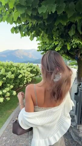 A woman with long brown hair wearing a light-colored slip dress and a white cardigan is walking along a cobblestone path next to a large bush of white flowers and trees. In the background, there are mountains and a body of water. The woman turns to face the camera and smiles, touching her hair. She is carrying a brown handbag.