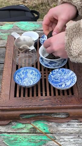 A pair of hands is shown breaking apart a chunk of dried tea leaves and placing them into a white ceramic gaiwan with a blue dragon pattern. The gaiwan sits on a matching saucer atop a wooden tea table. The table is slotted, with a handle, and has traces of peeling green paint. To the left of the gaiwan is a clear glass pitcher containing a metal strainer. Above and to the right of the gaiwan are several small white teacups, and underneath are two blue and white decorated plates. The scene appears to be outdoors in the daytime.